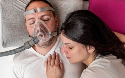 Hombre usando CPAP de viaje mientras duerme, acompañado de su pareja, ilustrando la continuidad del tratamiento al dormir fuera de casa.