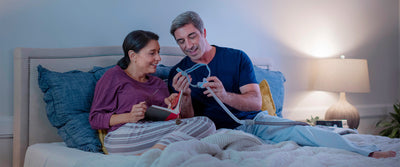 Una pareja recostada en la cama, ella sostiene un libro y él le muestra una mascarilla CPAP. La imagen ilustra la importancia de reemplazar los suministros del CPAP.