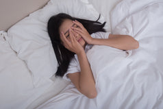 Mujer joven acostada en su cama cubriéndose el rostro con las manos debido a un intenso dolor de cabeza al despertar.