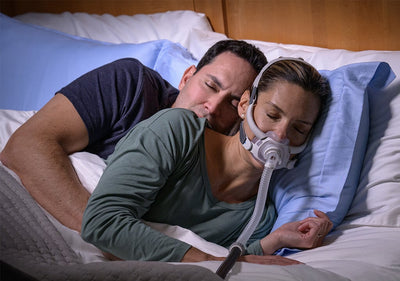Una pareja duerme en una cama. La mujer tiene una mascarilla y tubo de CPAP conectados, lo que sugiere que está recibiendo terapia para la apnea del sueño.