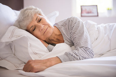  Mujer mayor duerme de lado en una cama con sábanas blancas. La habitación se ve iluminada. La imagen ilustra los ciclos de sueño. 