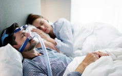Hombre durmiendo con una máquina CPAP para tratar la apnea del sueño, mientras su pareja descansa a su lado en la cama
