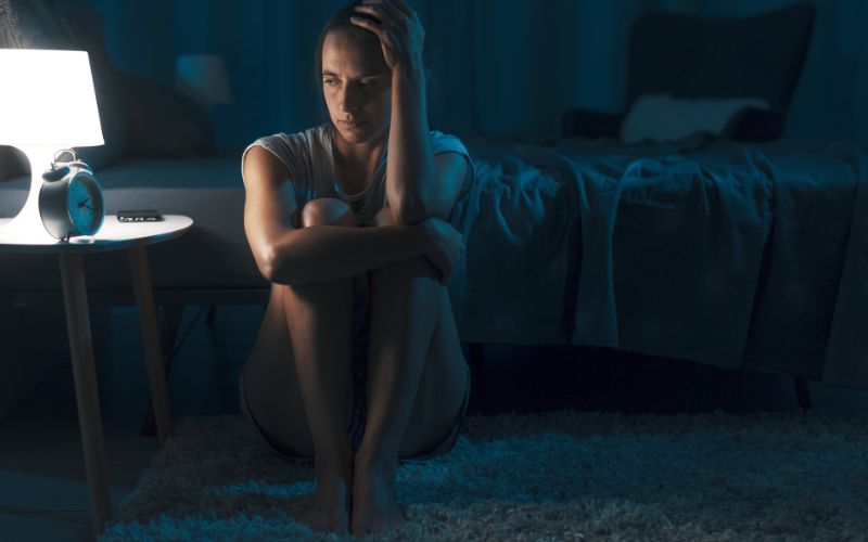 Mujer sentada en el suelo con insomnio y rostro de agotamiento, muestra efectos de la privación del sueño en cuerpo y mente.