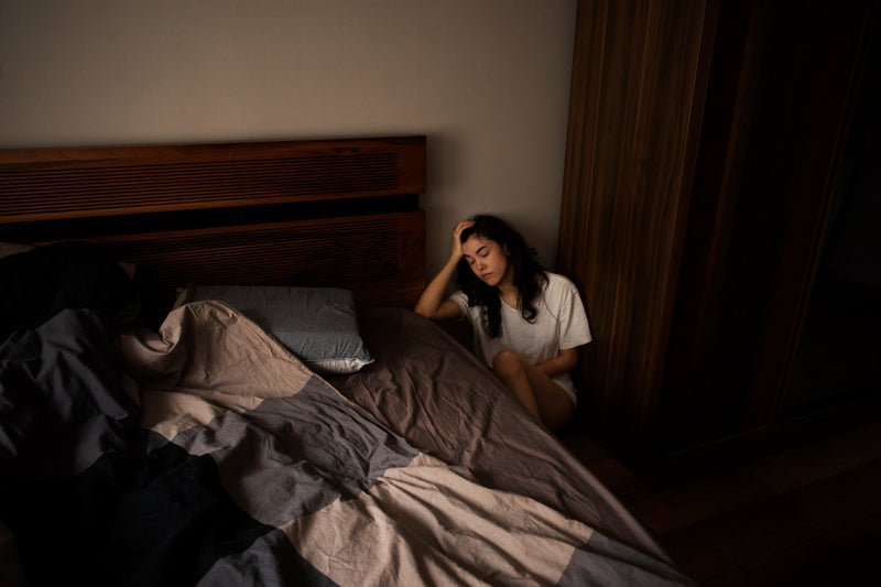 Una mujer joven de cabello largo está sentada en una esquina de su habitación, en penumbras, a un lado de su cama. Tiene una posición en la que parece dormitar sentada. 