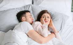 Hombre durmiendo y roncando mientras su pareja, con expresión molesta, se tapa los oídos en la cama debido al ruido.