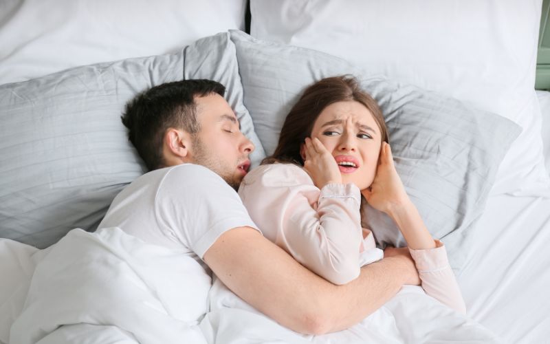 Hombre durmiendo y roncando mientras su pareja, con expresión molesta, se tapa los oídos en la cama debido al ruido.
