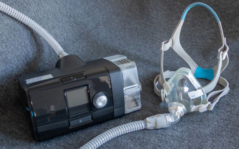 Equipo CPAP con humidificador integrado y mascarilla completa para terapia de apnea del sueño sobre superficie gris.