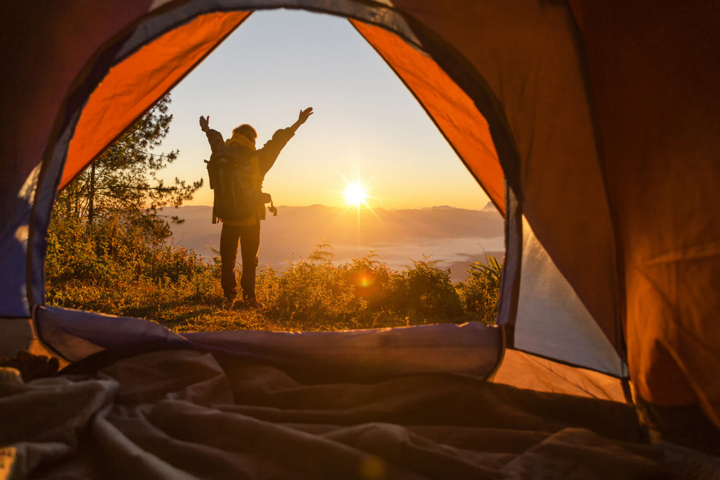 Toma desde el interior de una tienda de campaña; se ve a un hombre que ve la salida del sol desde una montaña y extiende sus brazos en señal de disfrute del paisaje. 