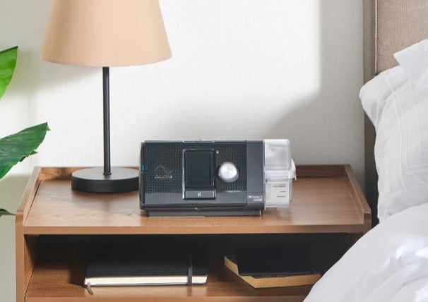 Equipo CPAP con humidificador integrado sobre un buró, al lado de una cama, listo para usarse durante la noche.