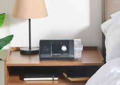 Equipo CPAP con humidificador integrado sobre un buró, al lado de una cama, listo para usarse durante la noche.